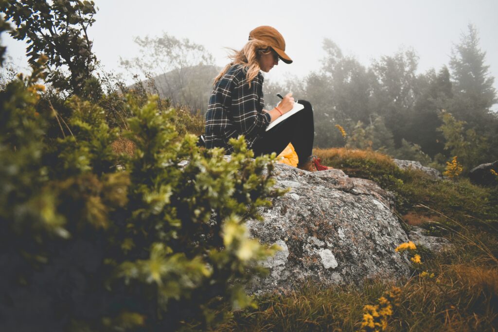 An image of a young woman wearing a ball cap, sitting on a rock and writing in a notebook while surrounded by nature