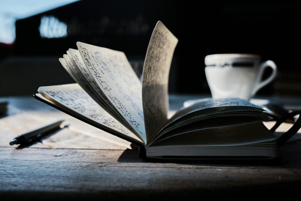 An image of an open notebook with lots of writing on its pages. It's sitting on a wooden surface with a pen and a white mug nearby in front of a dark background.