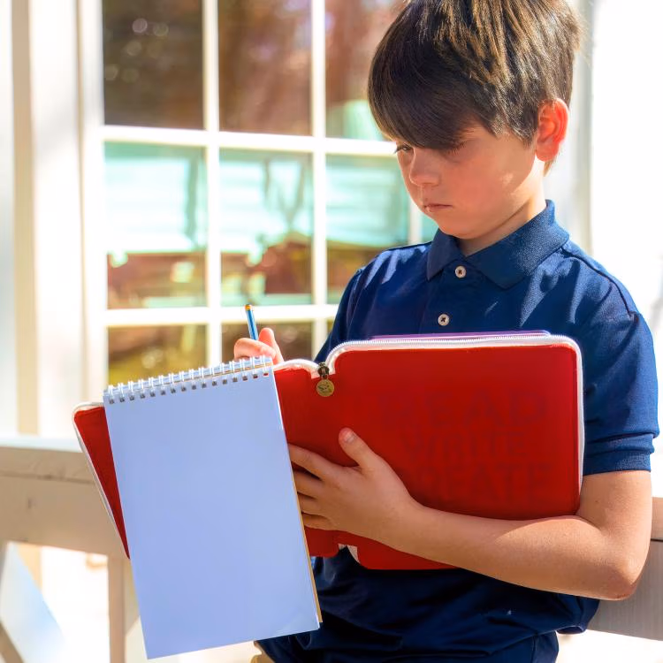 Young boy in a navy polo writes in a red notebook while holding a white spiral notebook outdoors by a window.